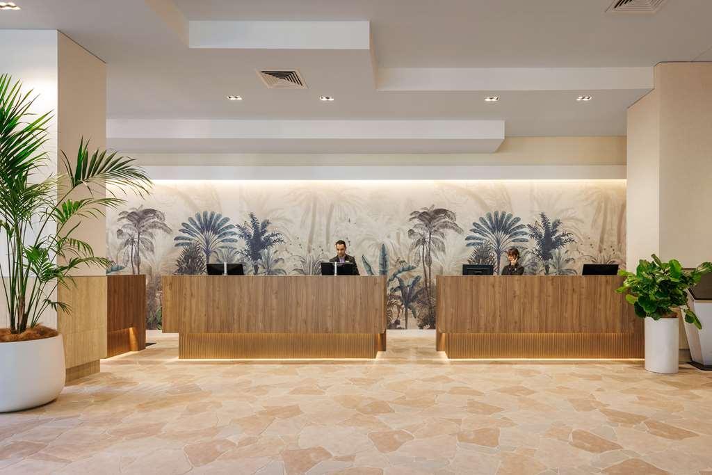 아모라 브리즈번 호텔 내부 사진 The photo shows a modern reception area with two wooden reception desks. Behind the desks, there is a decorative wall featuring a large mural of tropical plants and palm trees, creating a serene atmosphere. The space is well-lit with overhead lights,